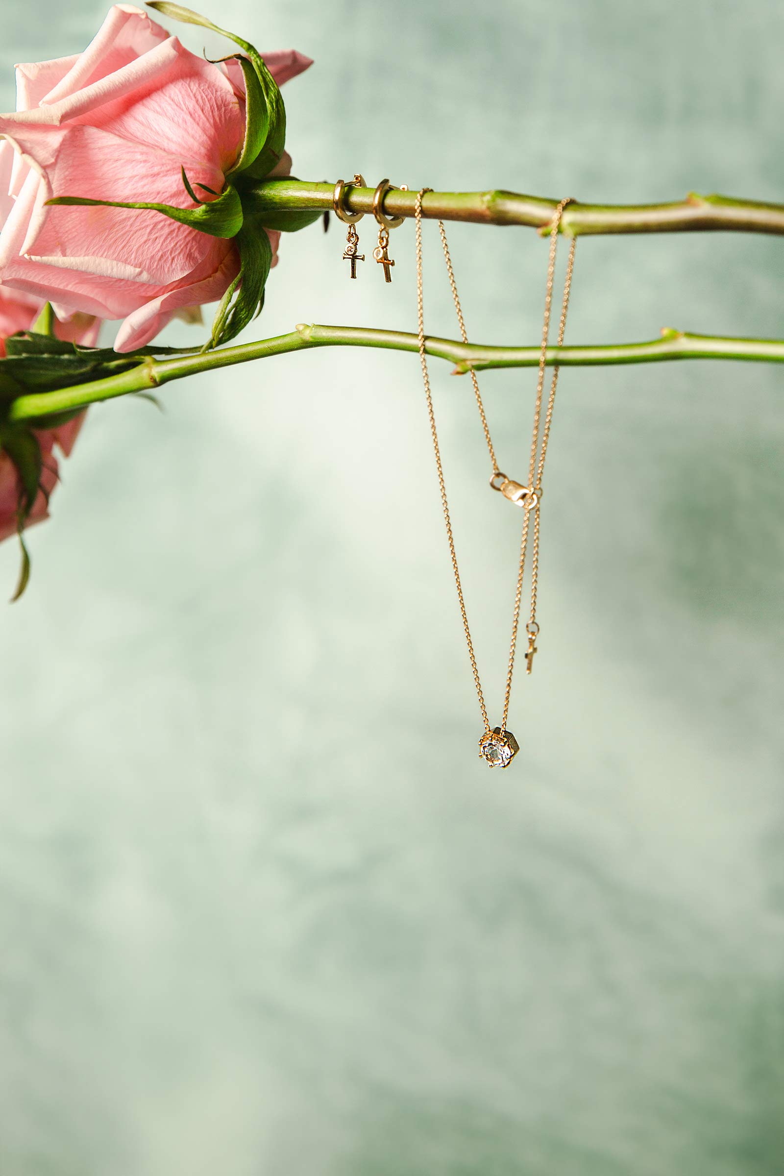 Rizen Jewelry Chispa Collection featuring the 18k gold vermeil Ebenezer white topaz solitaire necklace and Calvary collection Cross Huggie Hoop Earrings on a pink rose stem. 