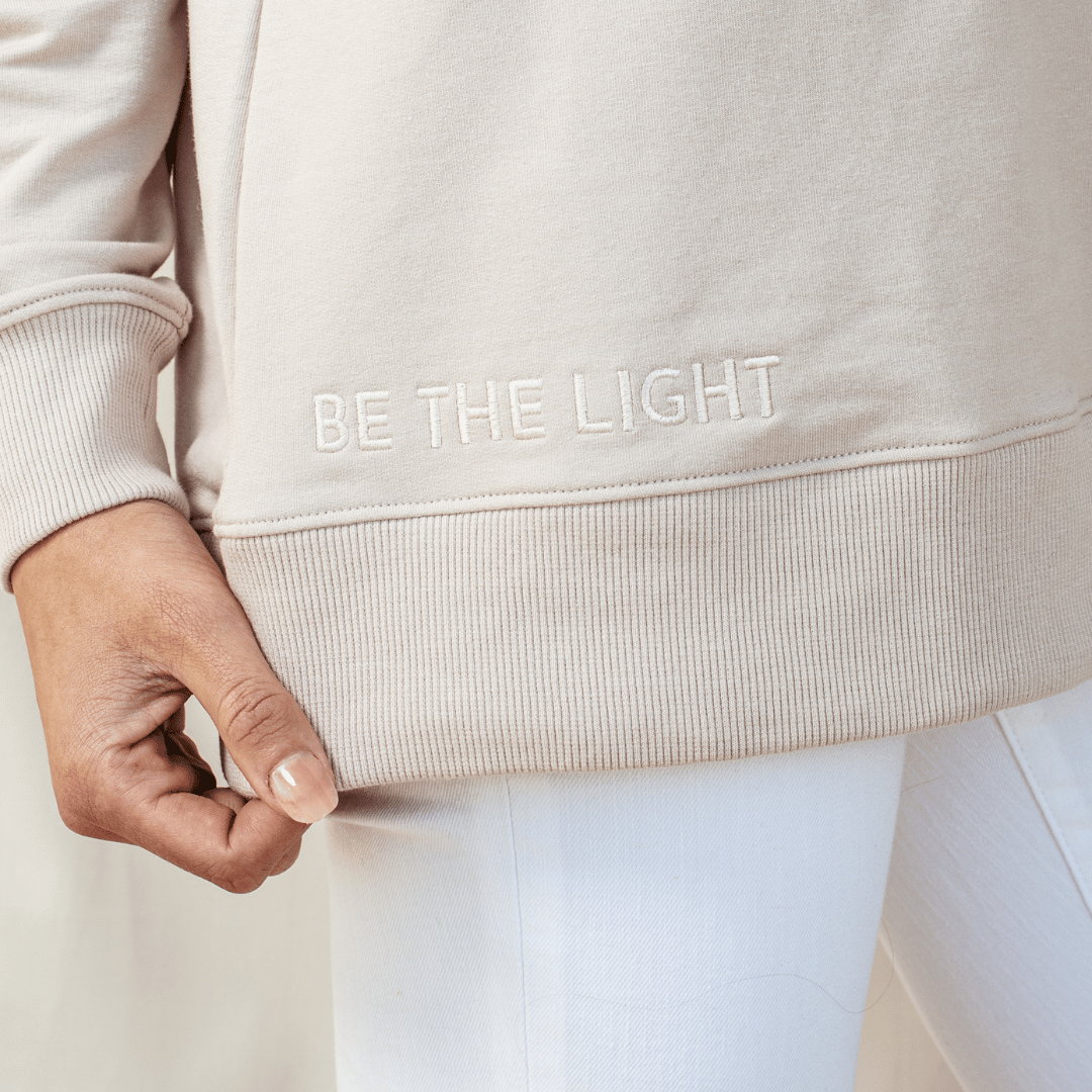 Close up of "BE THE LIGHT" embroidery on the shore beige "Becoming. Philippians 1:6" Peruvian cotton crew sweatshirt from the Be The Light Collection by Rizen Jewelry.