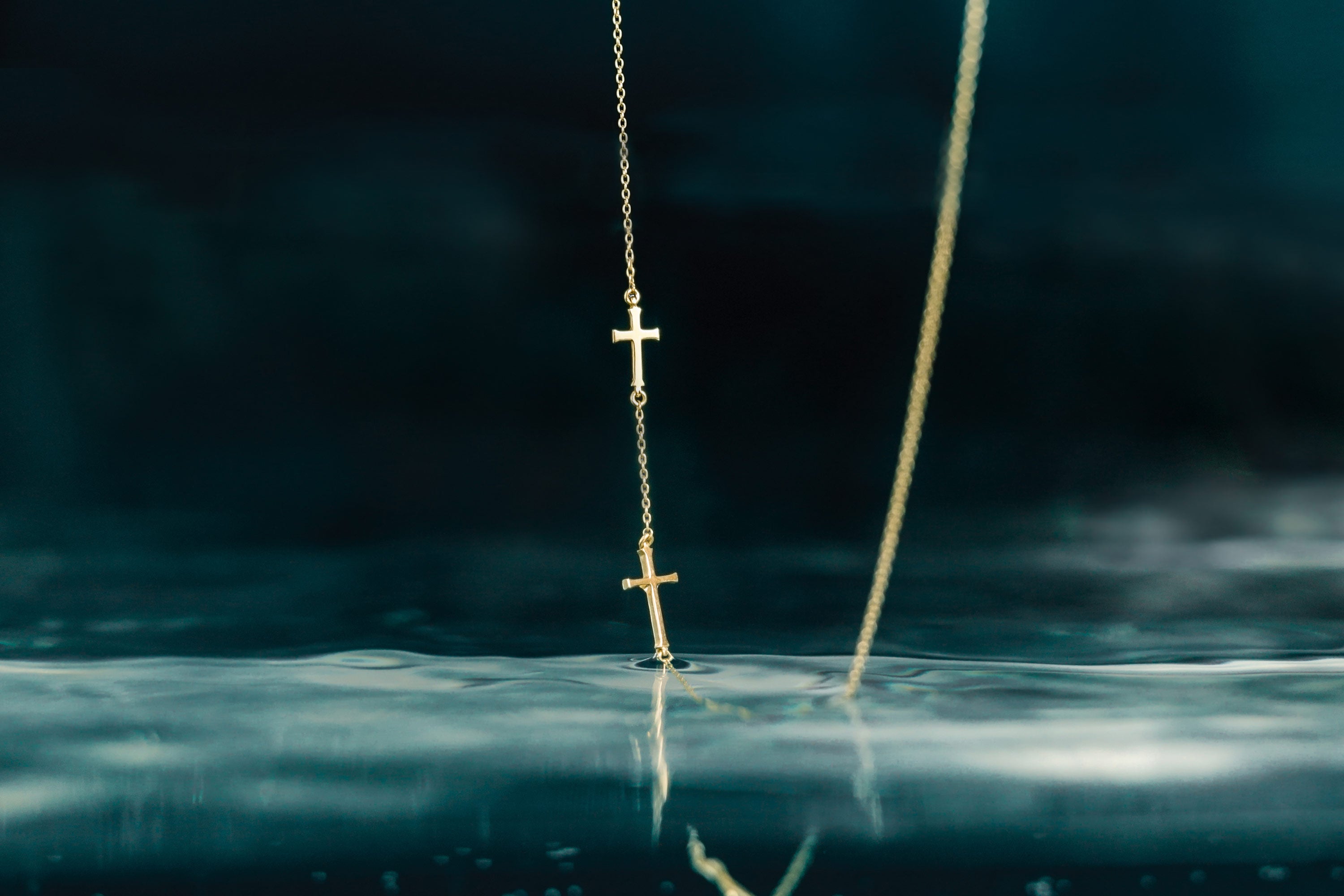 Rizen Jewelry Calvary triple cross necklace in 18kt yellow gold necklace dripping into deep water.