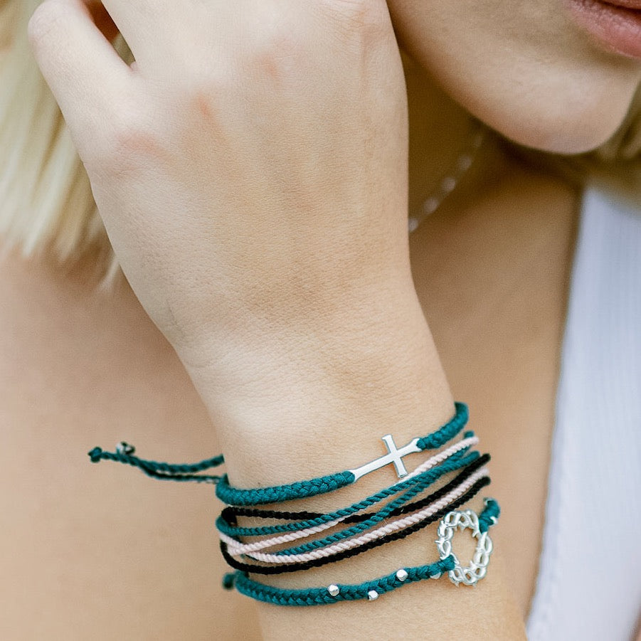 Christian model wearing silver cross bracelet engraved "STAY SALTY" hand braided azure blue cotton cords by Rizen Jewelry Made 4 Ministries collection. Layered with beaded crown of thorns and multi cord bracelets.