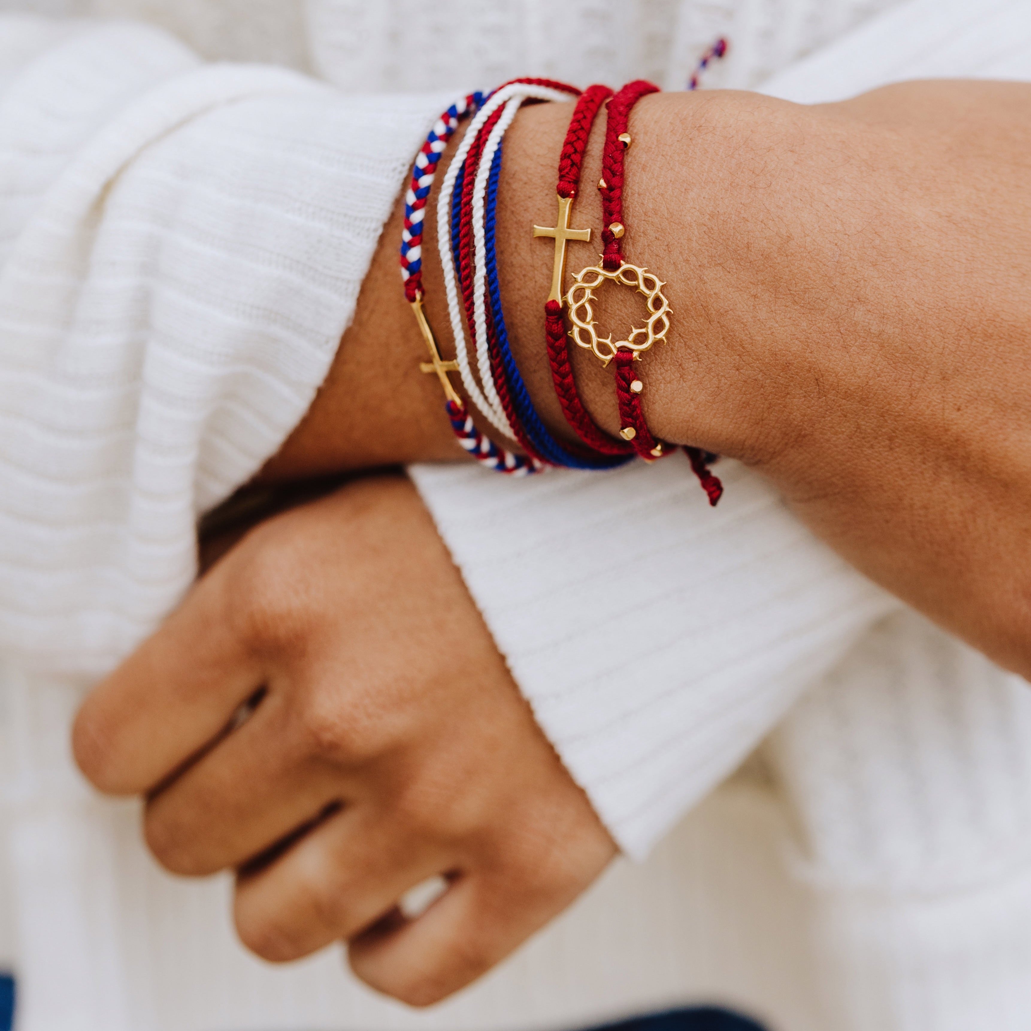 Christian model wearing Rizen Jewelry Made 4 Ministries gold We Trust in God Bracelet Set. Featuring Red Garnet Beaded Crown of Thorns; Red, White, and Blue We Trust in God Cross bracelet; Garnet Red STAY SALTY braided bracelet; and peaceful Red, White, and Blue Be Magnified multi-cord bracelet.