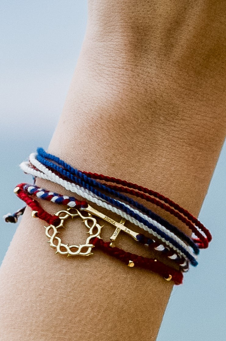 christian model wearing gold cross bracelet hand braided in red, white, blue cotton cord, engraved "WE TRUST IN GOD" with Rizen Jewelry and Made 4 Ministries round disc tag. Layered with garnet red beaded crown of thorns and red white blue multi cord bracelet 
