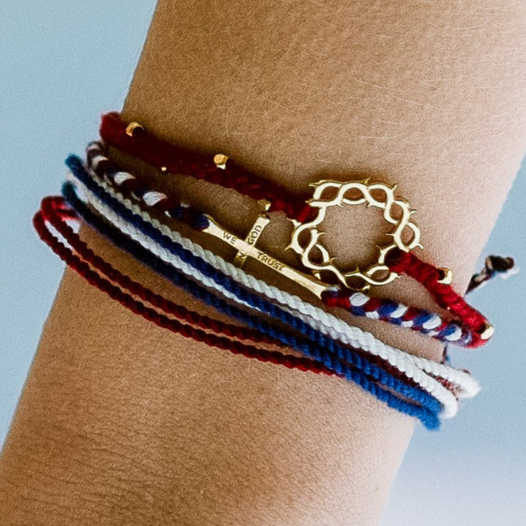 christian model wearing gold cross bracelet hand braided in red, white, blue cotton cord, engraved "WE TRUST IN GOD" with Rizen Jewelry and Made 4 Ministries round disc tag. Layered with garnet red beaded crown of thorns and red white blue multi cord bracelet