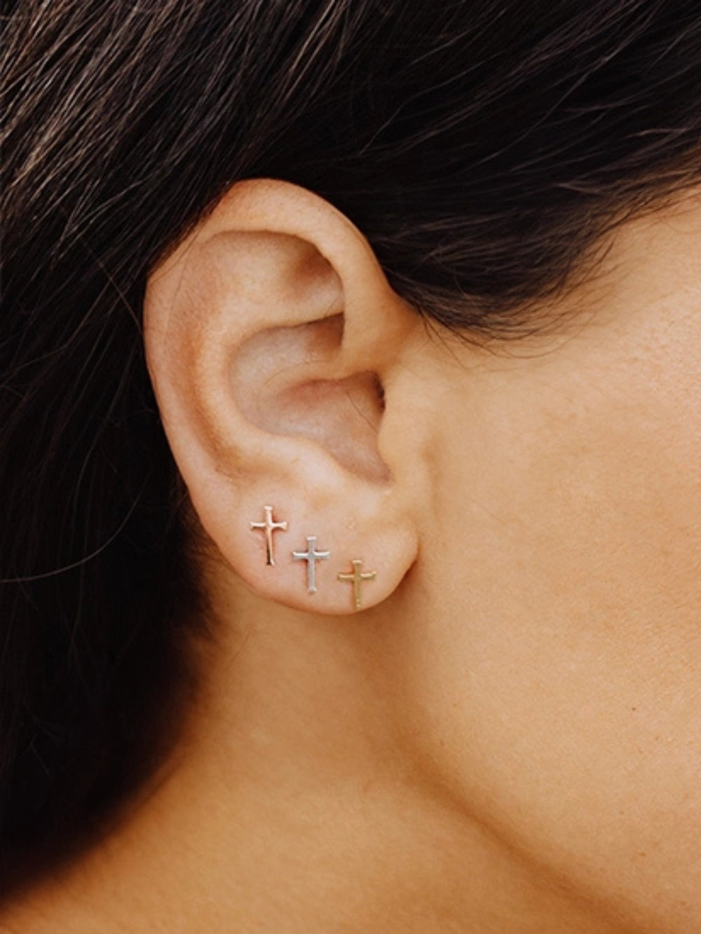 Close up of Christian woman wearing 3 pairs of the mini Cross Stud Earrings in sterling silver, rose, and yellow gold plated from the Calvary Collection by Rizen Jewelry.