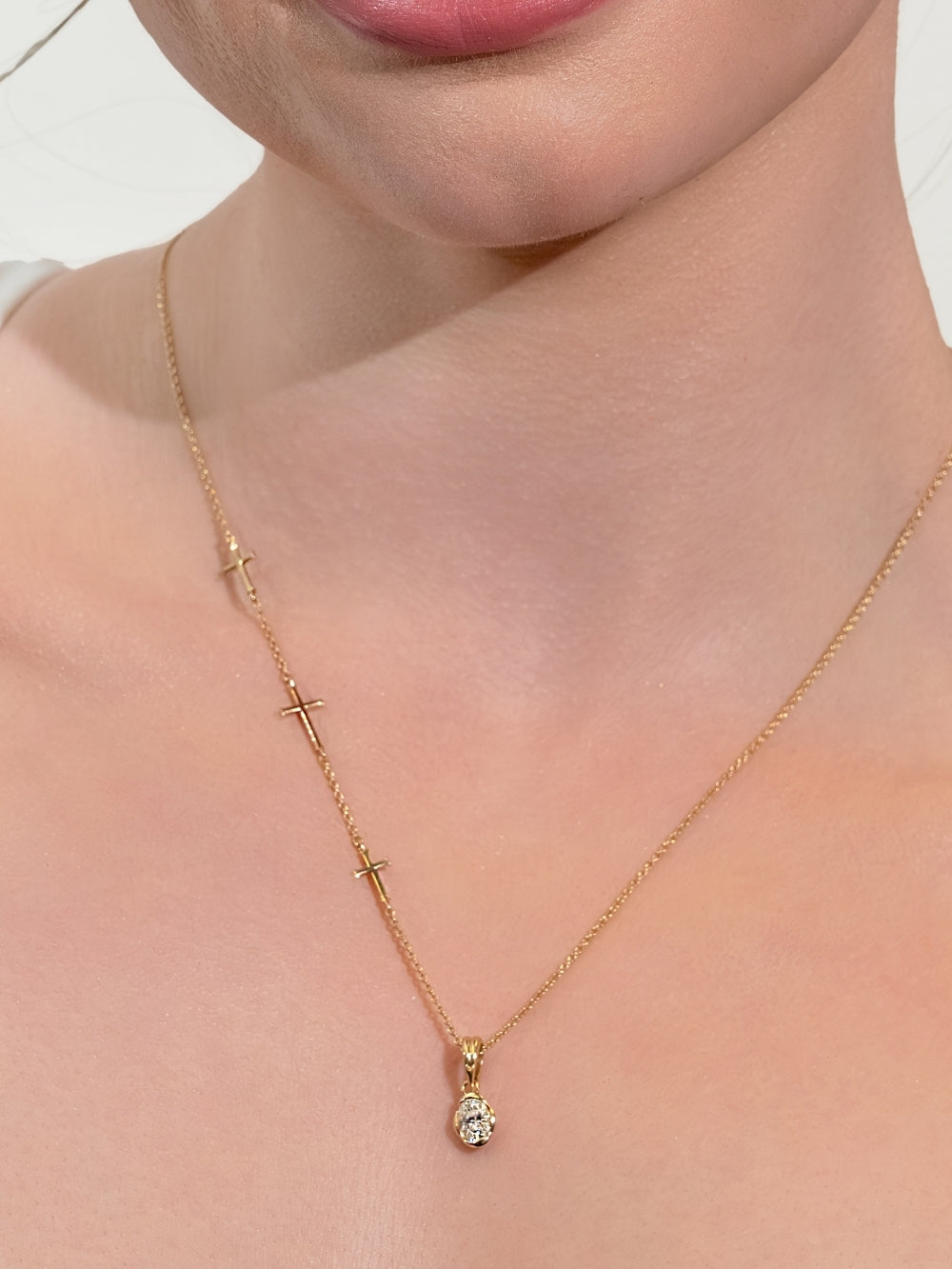 Women's Christian jewelry model wears the Calvary three-cross necklace styled with the White Sapphire Fully Known Prayer Pendant by Rizen Jewelry features A radiant oval brilliant-cut white sapphire pendant, reflecting innocence, our new identity, and the luminous pure heart of April, cradled gently in refined 18k gold plating over sterling silver. 