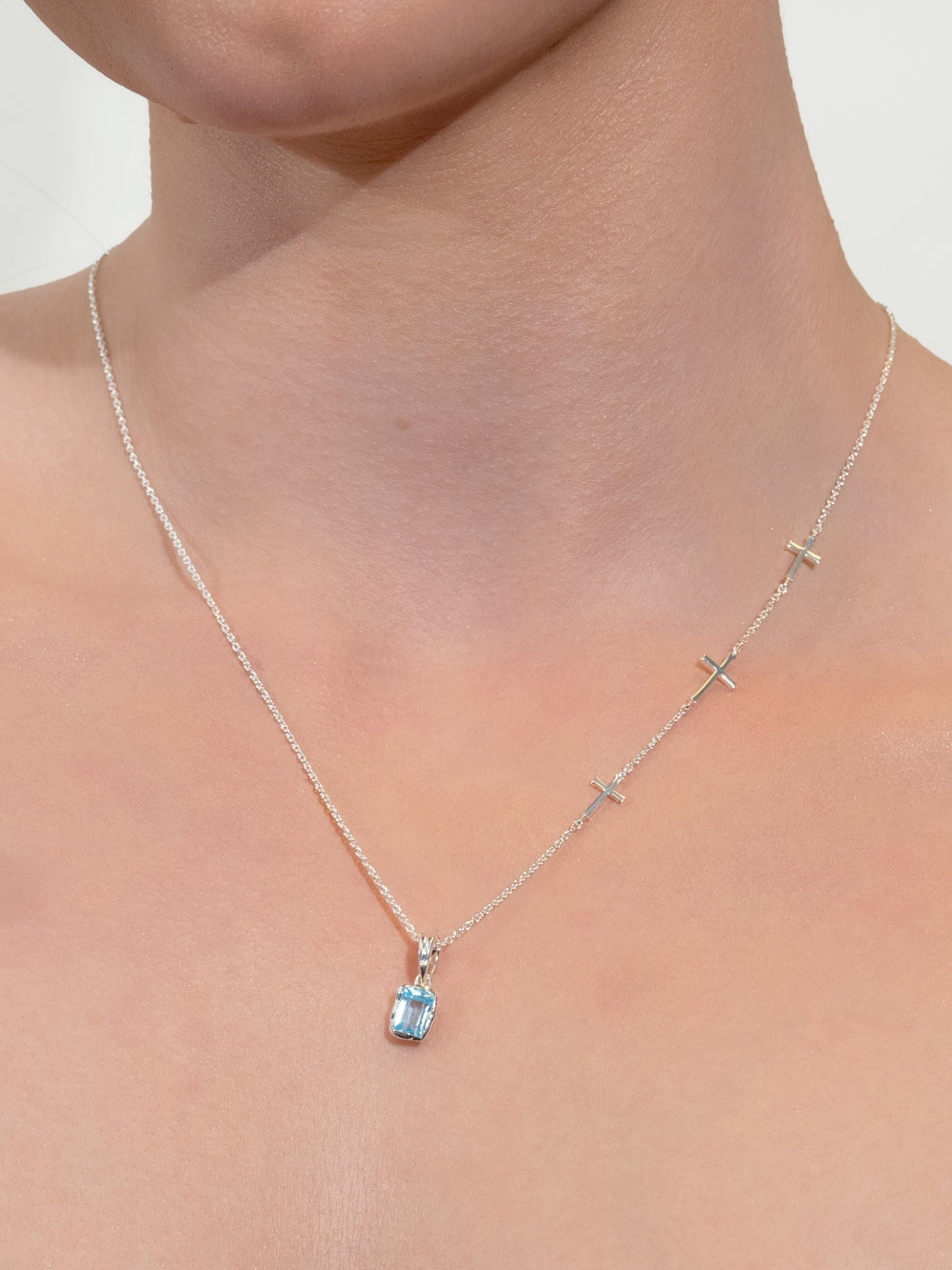 Women's Christian jewelry model wearing the Calvary three-cross necklace with an added Sky Blue Topaz fully known prayer pendant.