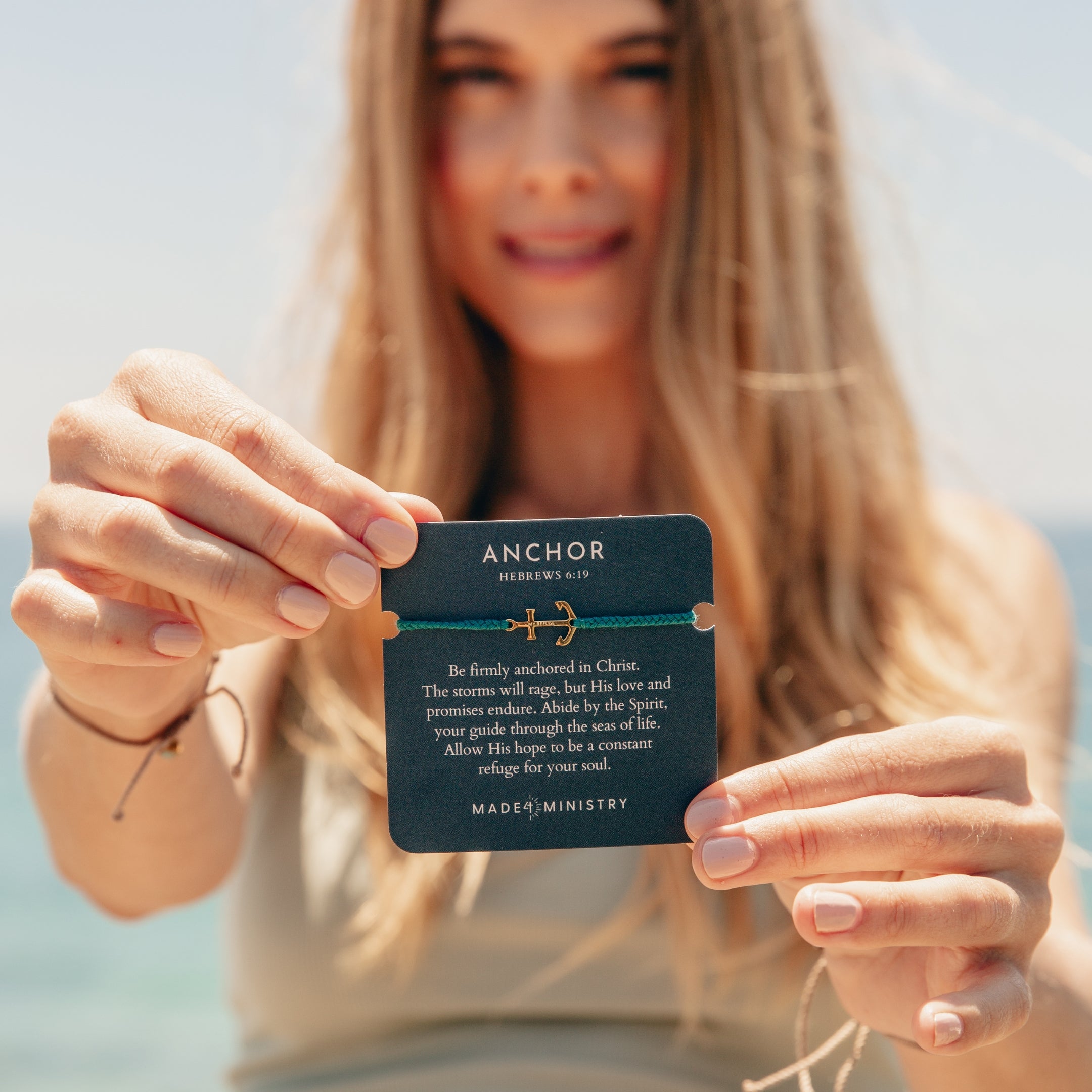 Classic nautical anchor bracelet, delicately engraved with the inscription “MY REFUGE,” hand-braided from naturally dyed azure blue cotton cord. Available in 18k gold or sterling silver finish from the Made4Ministry Collection by Rizen Jewelry. Held on devotion card by female women christian jewelry model. 