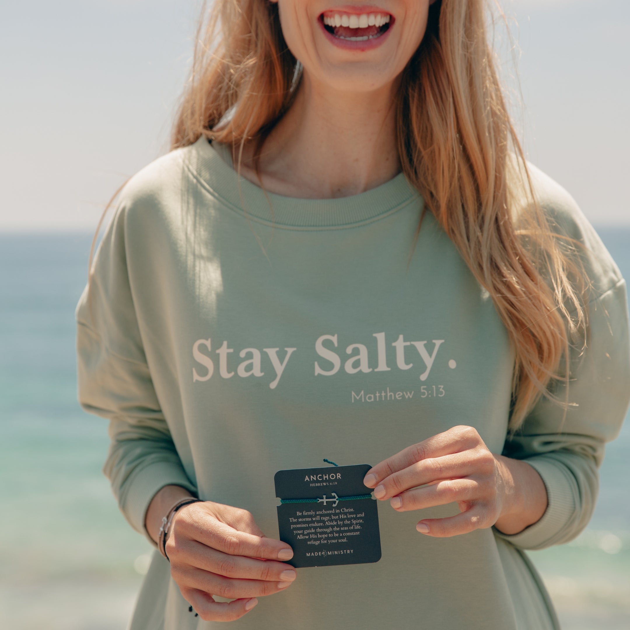 Women's Christian Jewelry model wearing the Be The Light Stay Salty Crew Sweatshirt holding the Anchor Bracelet featured on the devotion card. 