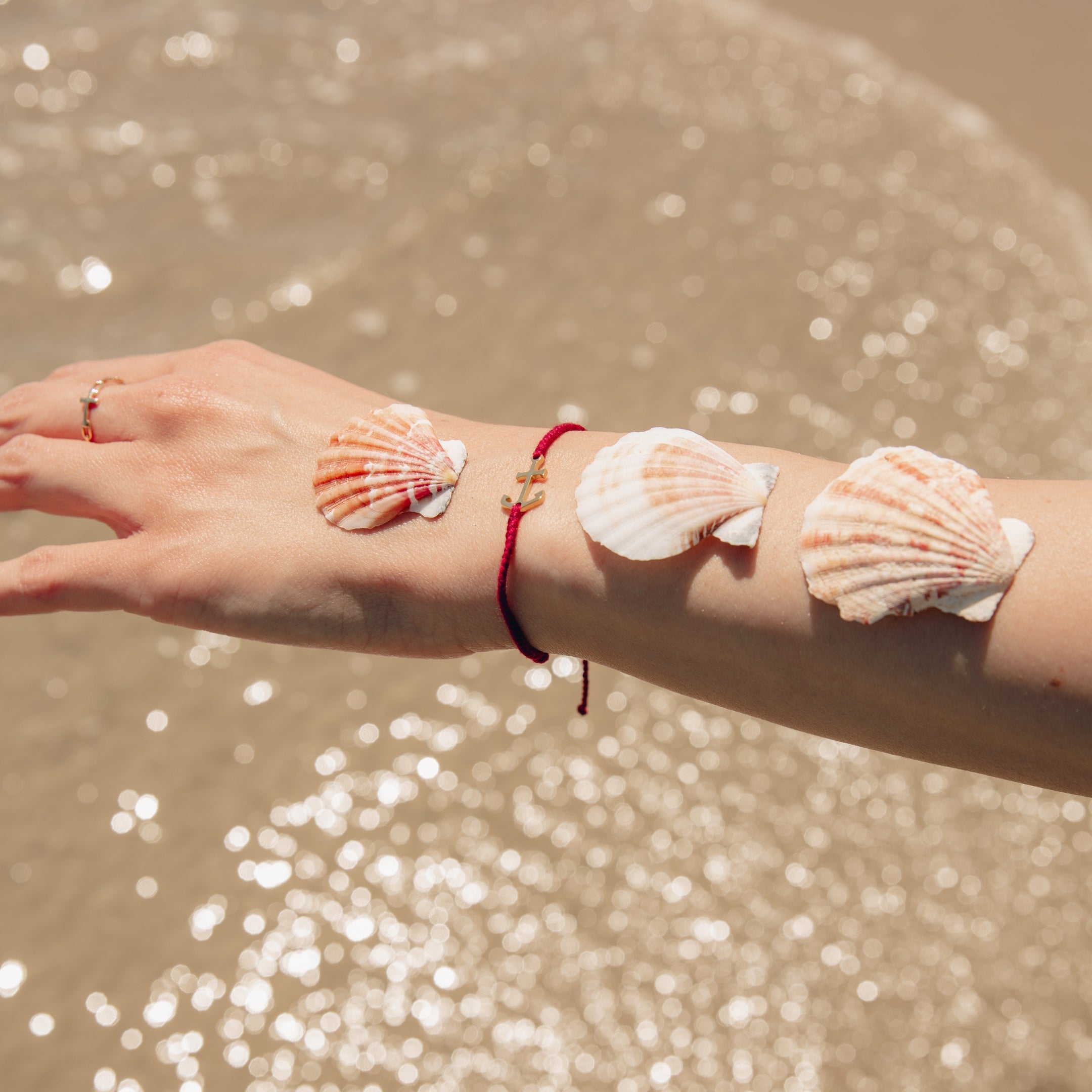 Womens christian jewelry model balancing 3 scalloped shells and the Classic nautical anchor bracelet, delicately engraved with the inscription “MY REFUGE,” hand-braided from naturally dyed garnet red cotton cord. Available in 18k gold or sterling silver finish from the Made4Ministry Collection by Rizen Jewelry.