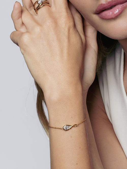 A close-up lifestyle shot of a Christian female model wearing a delicate gold chain bracelet. Rizen Jewelry's Solace Teardrop Bracelet has a tear drop shaped clear simulated diamond nesteled inside a flowing infinity symbol. She is also wering the matching ring.