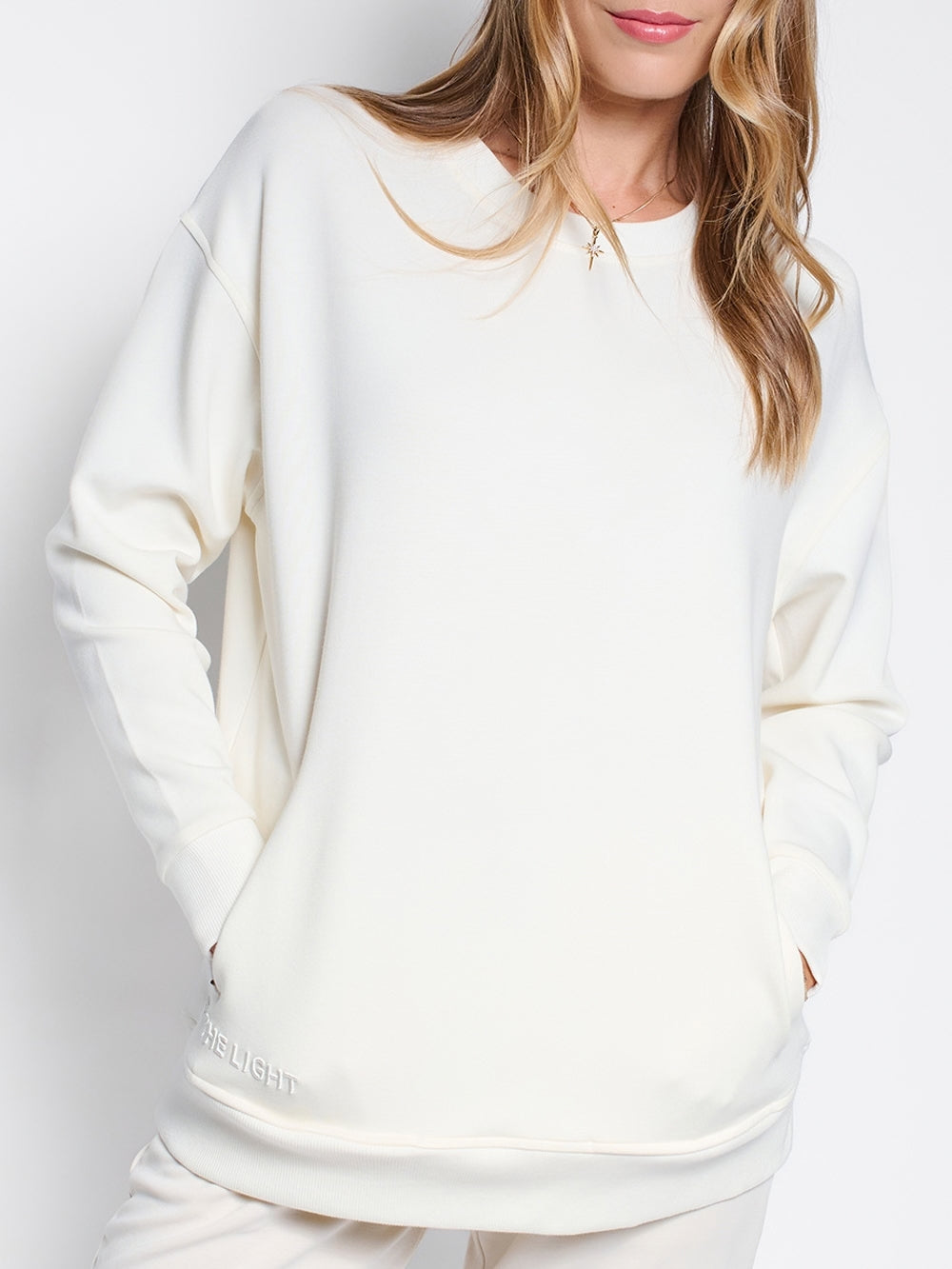 Christian female sweatshirt model wearing Joy crewneck sweatshirt with Be The Light embroidery and discreet front pockets with matching Adoring Star necklace