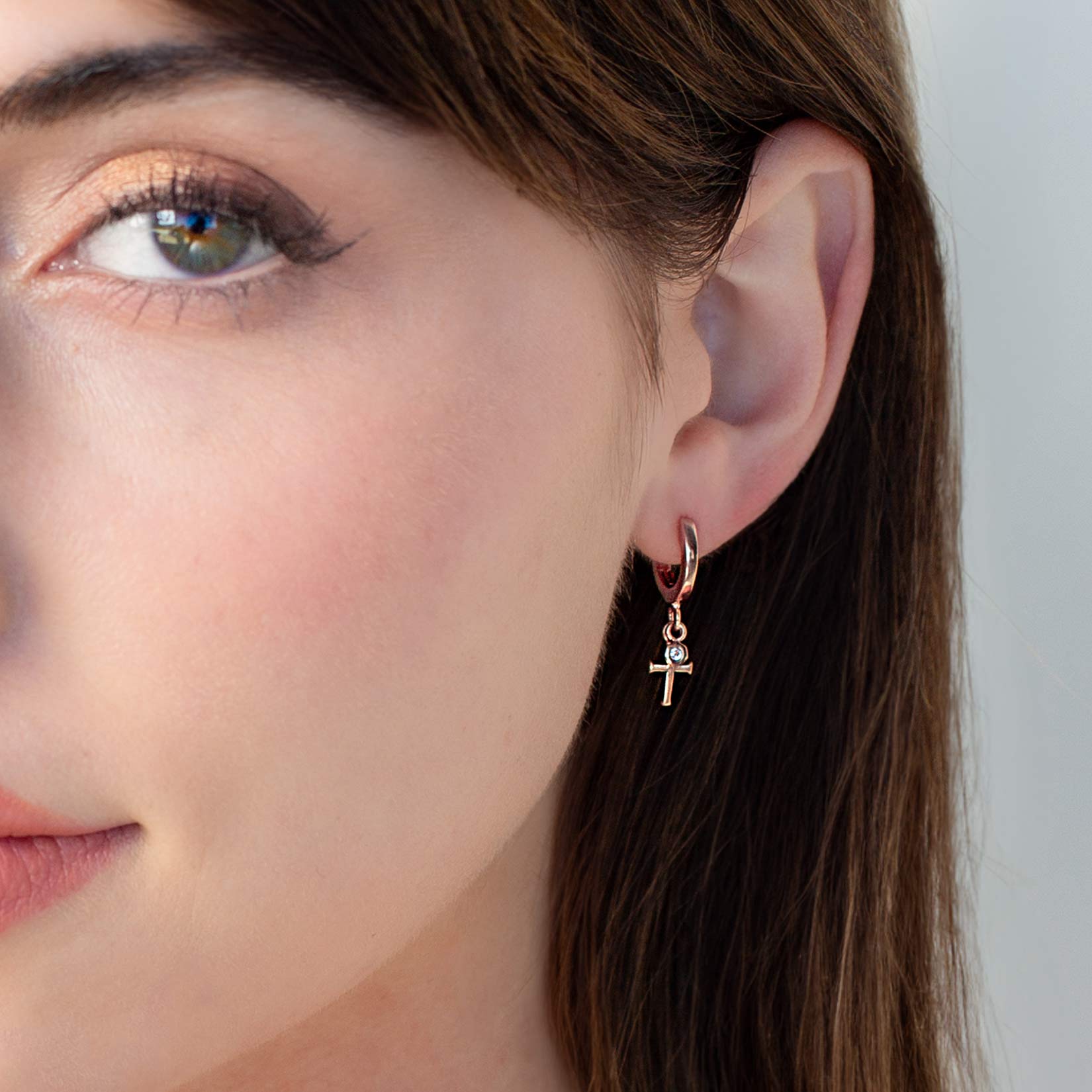 Close up of a Christian woman wearing the Calvary Huggie Hoop Earrings with white topaz stone dangle in 18k rose gold plated from the Calvary Collection. A timeless baptism gift by Rizen Jewelry.
