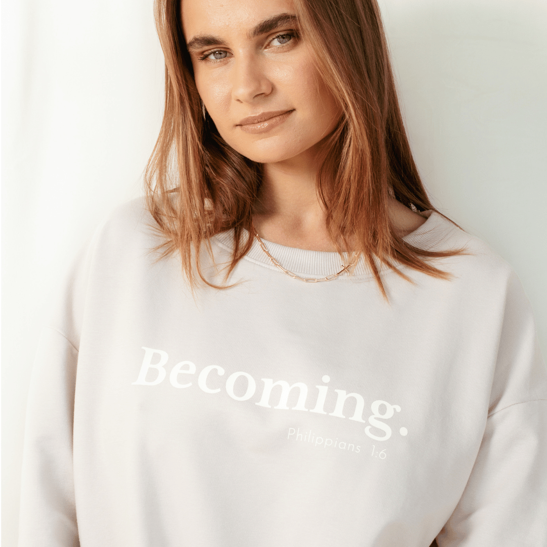 Christian woman wearing the shore beige "Becoming. Philippians 1:6" Peruvian cotton crew sweatshirt from the Be The Light Collection by Rizen Jewelry.