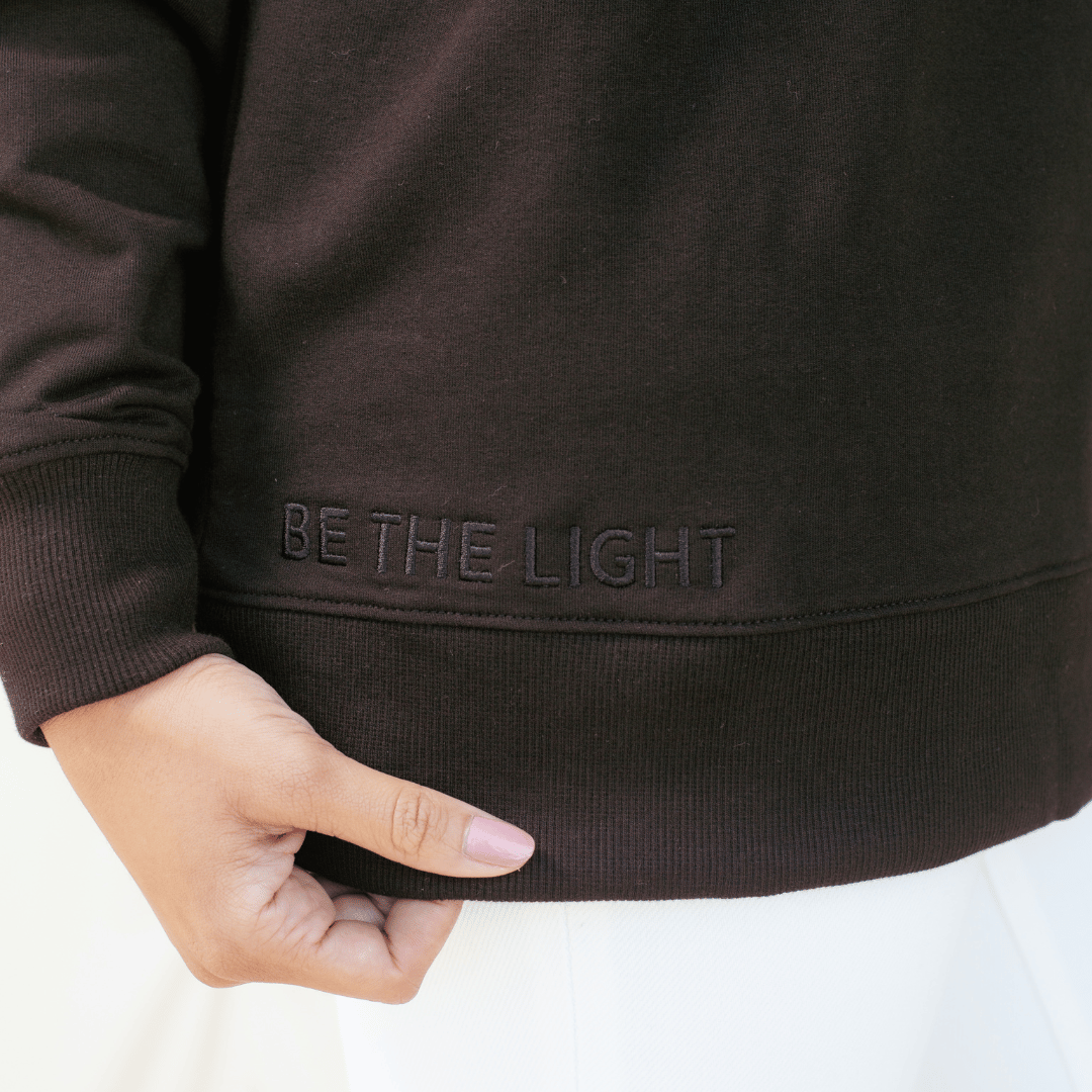 Close up of "BE THE LIGHT" embroidery on the black " Loved." sweatshirt from the Be The Light Collection by Rizen Jewelry.