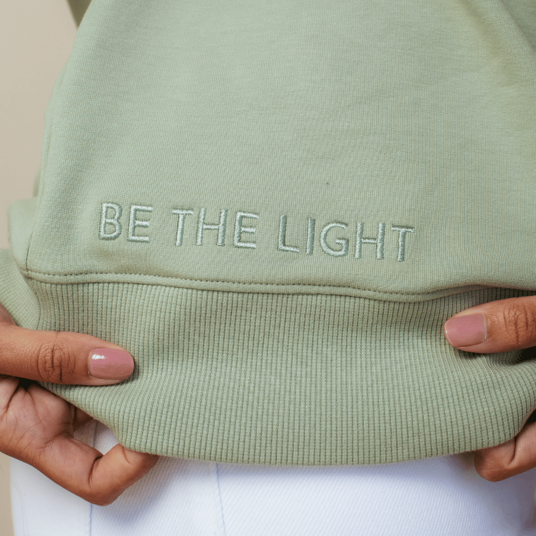 Close up of "BE THE LIGHT" embroidery above the hem on the wave green "Stay Salty. Matthew 5:13" Peruvian cotton crew sweatshirt from the Be The Light Collection by Rizen Jewelry.