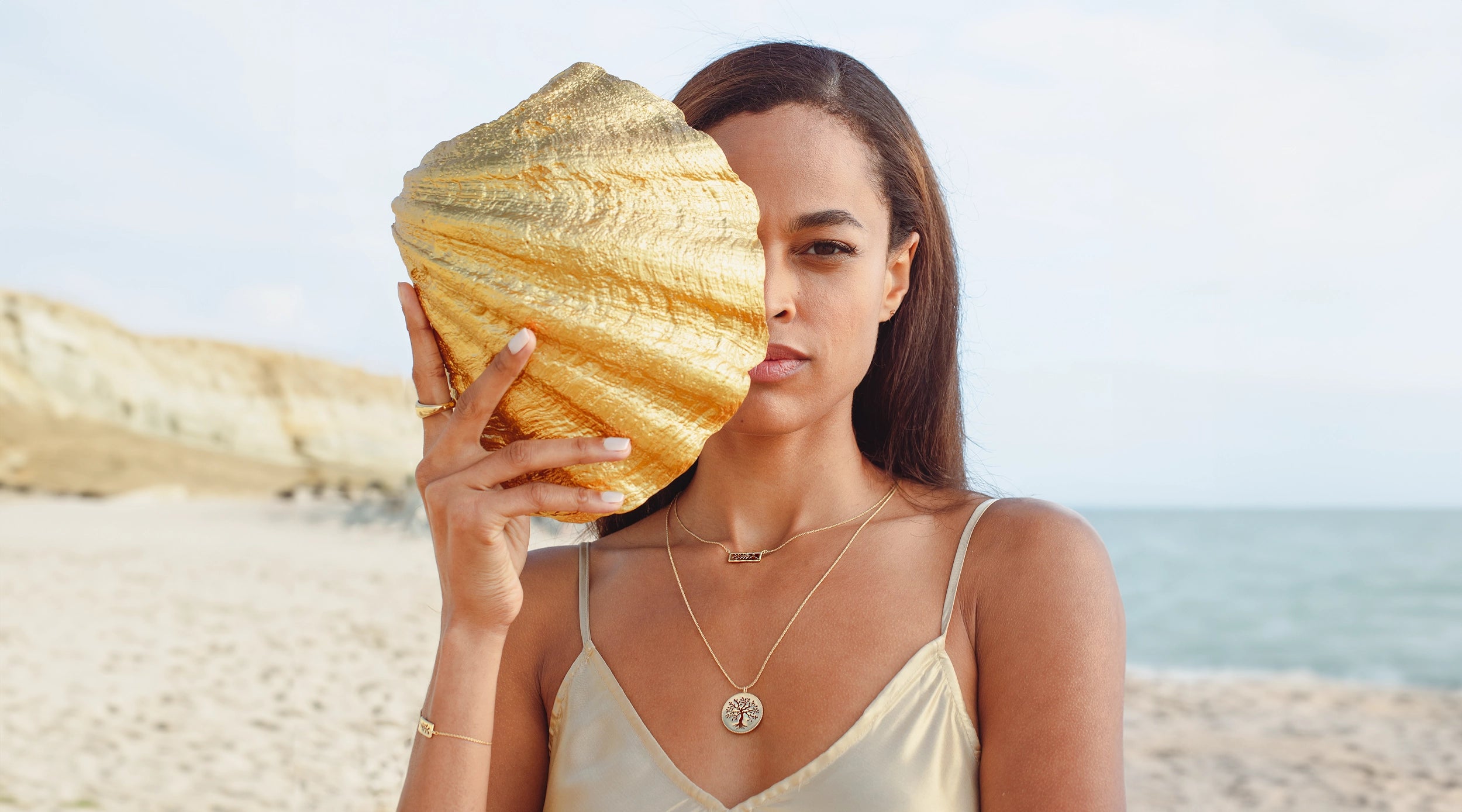 Christian woman holding gold shell wearing the gold Rooted Collection by Rizen Jewelry.