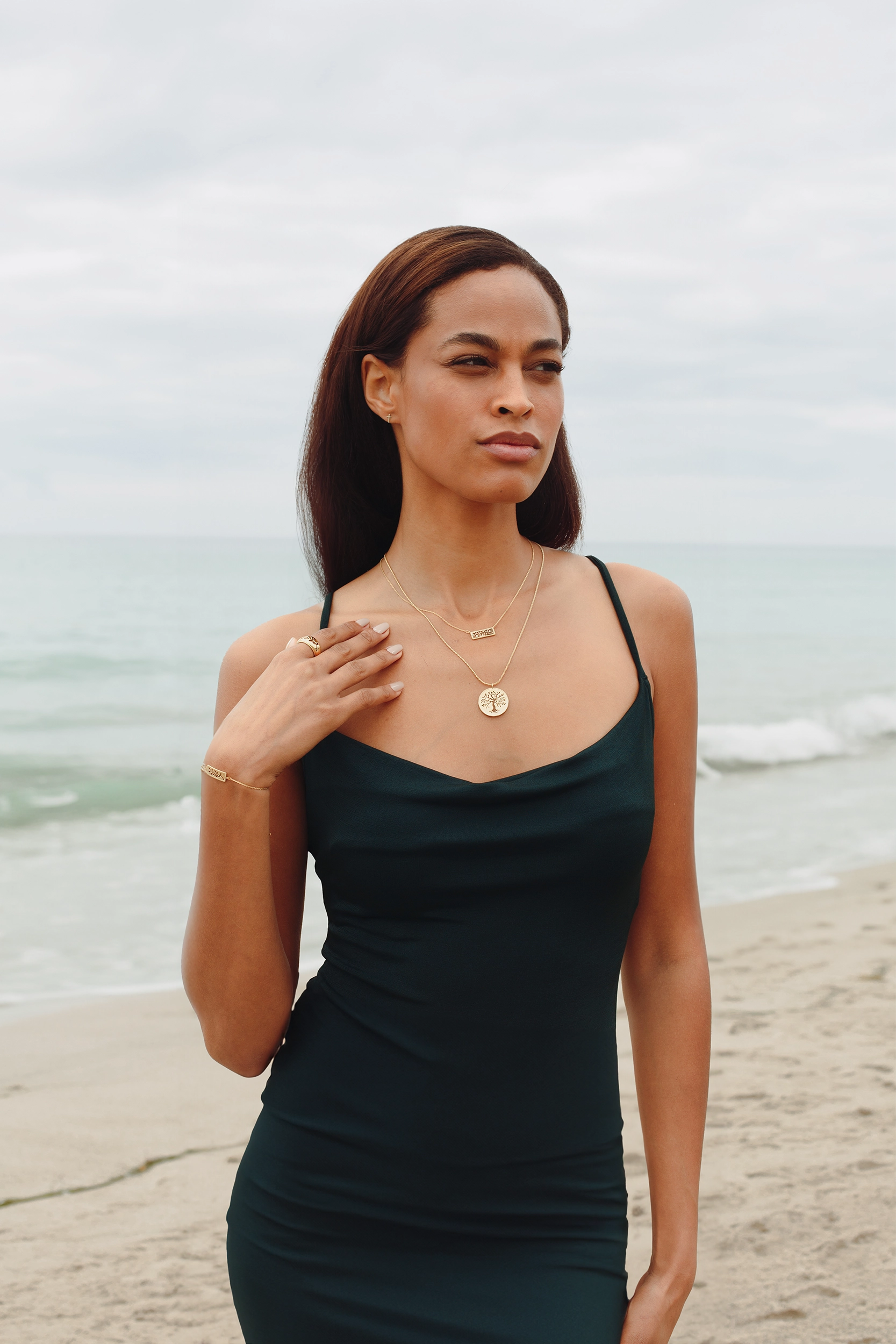Woman at beach wearing Rizen Jewelry Rooted Collection in 18kt yellow gold vermeil, includes the branch ring, bracelet and necklace, olive tree necklace and mini cross stud earring. 