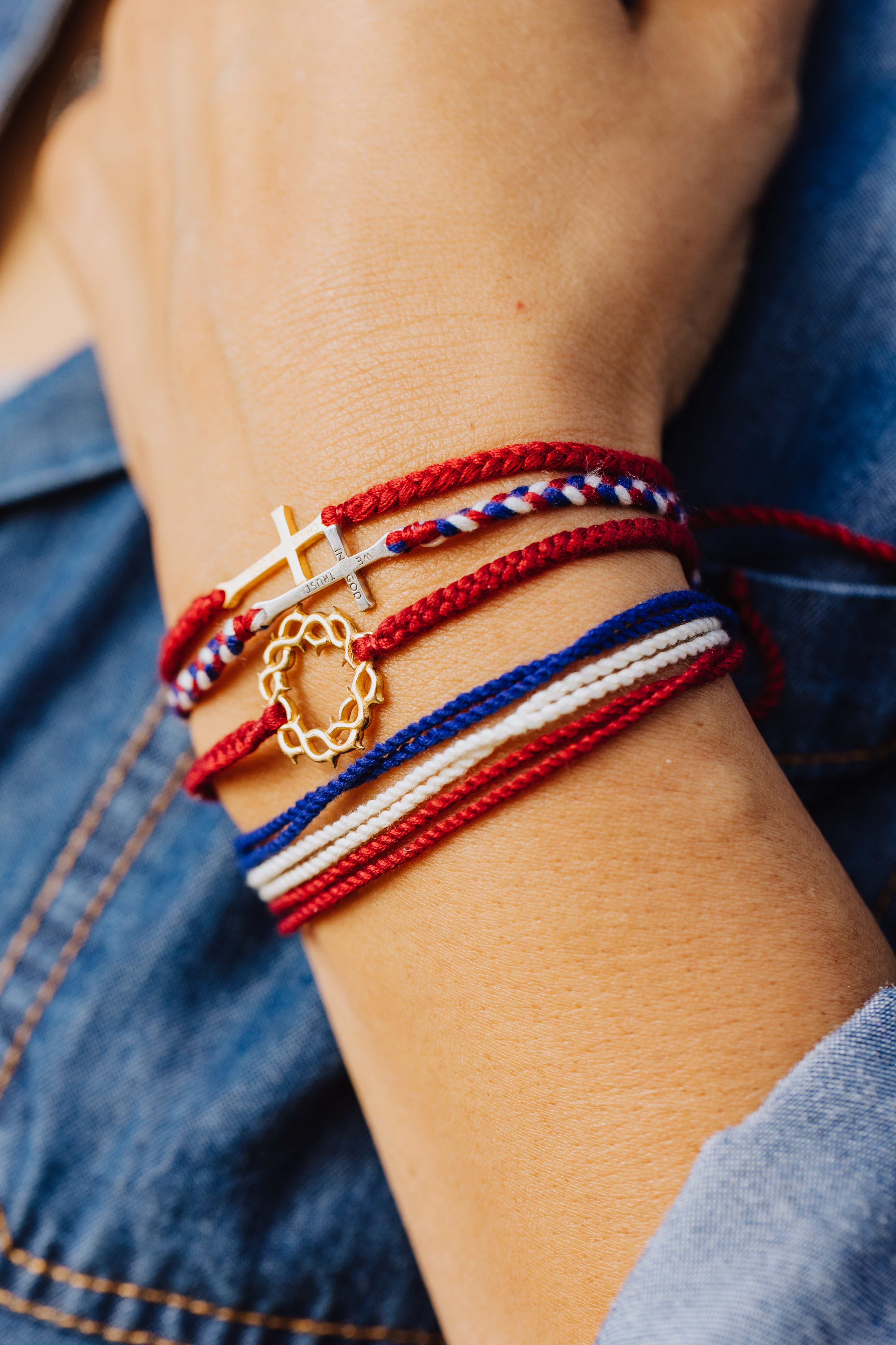 Christian model wearing silver  cross bracelet hand braided in red, white, blue cotton cord, engraved "WE TRUST IN GOD" with Rizen Jewelry and Made 4 Ministries round disc tag. Layered with multiple red white and blue Made 4 Ministries bracelets. 