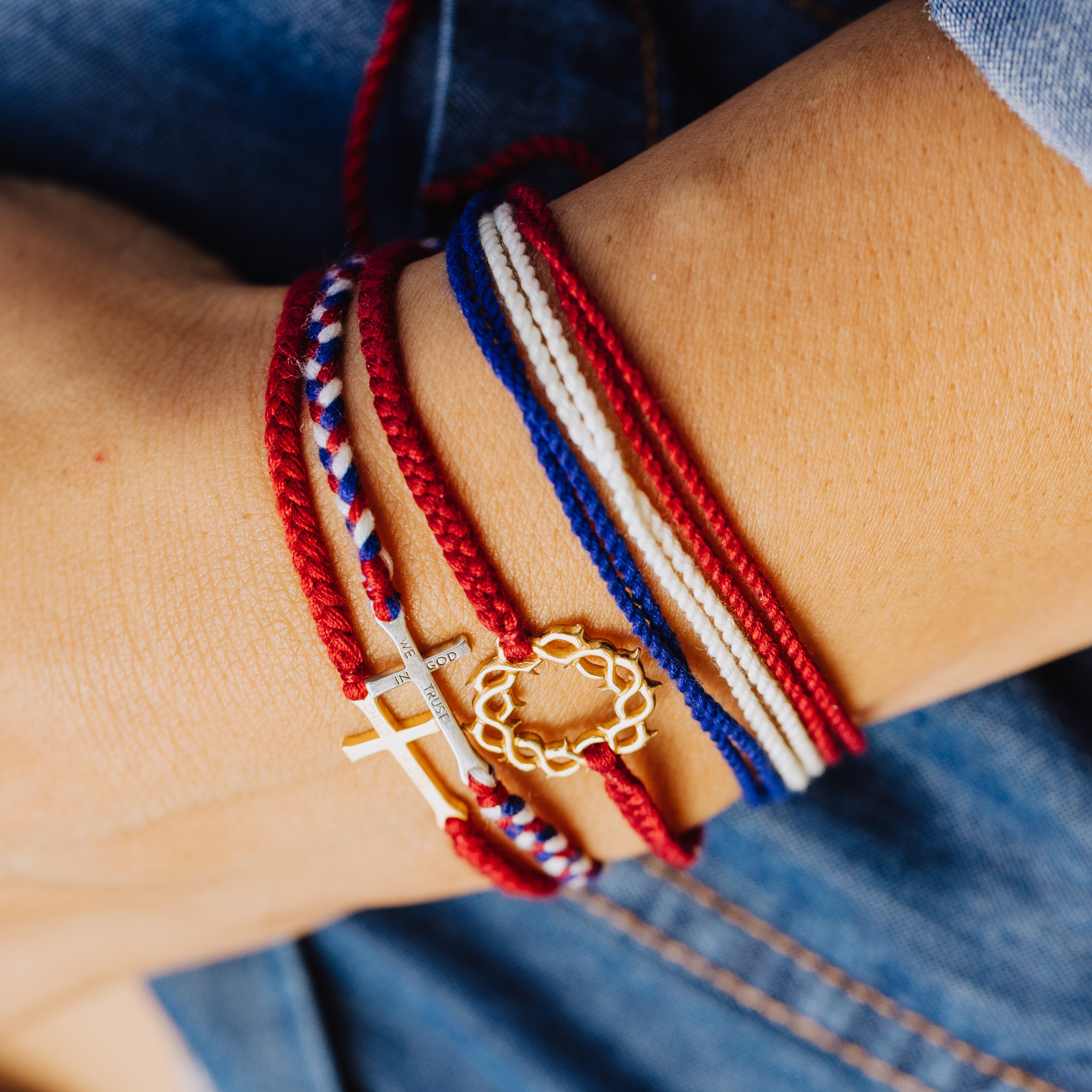 Christian model wearing silver  cross bracelet hand braided in red, white, blue cotton cord, engraved "WE TRUST IN GOD" with Rizen Jewelry and Made 4 Ministries round disc tag. Layered with multiple red white and blue Made 4 Ministries bracelets. 