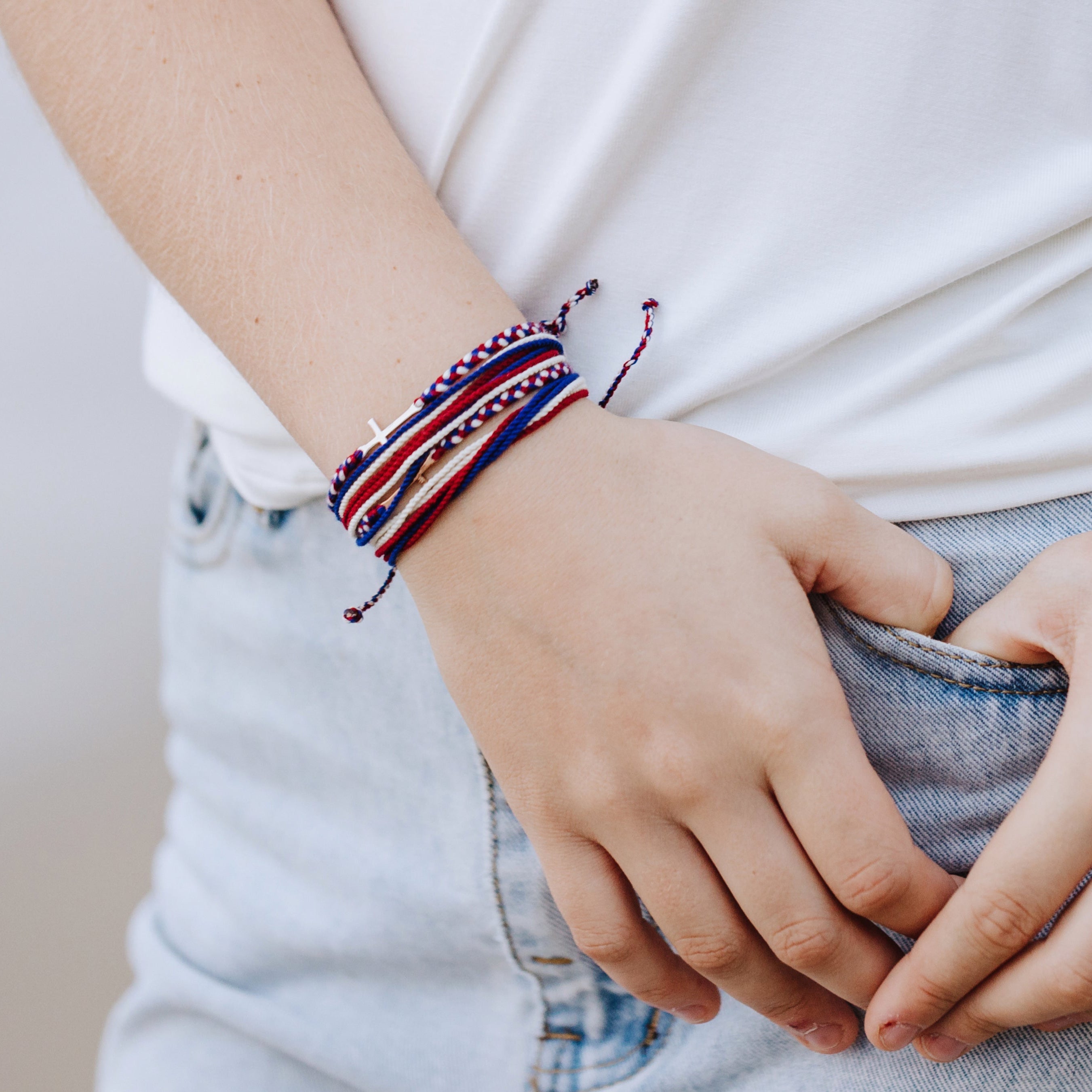 Christian model wearing rose gold cross bracelet hand braided in red, white, blue cotton cord, engraved "WE TRUST IN GOD" with Rizen Jewelry and Made 4 Ministries round disc tag. and multiple other red, white and blue cross bracelets. 